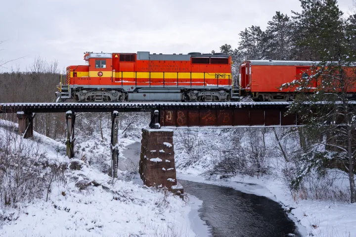 Red-orange train crossing snowy bridge over a narrow creek, surrounded by trees in winter.
