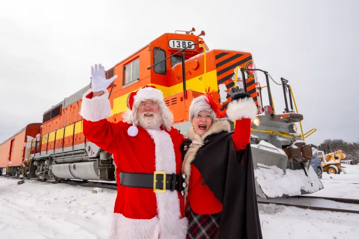 Santa and Mrs. Claus waving in front of a festive train in snowy landscape.