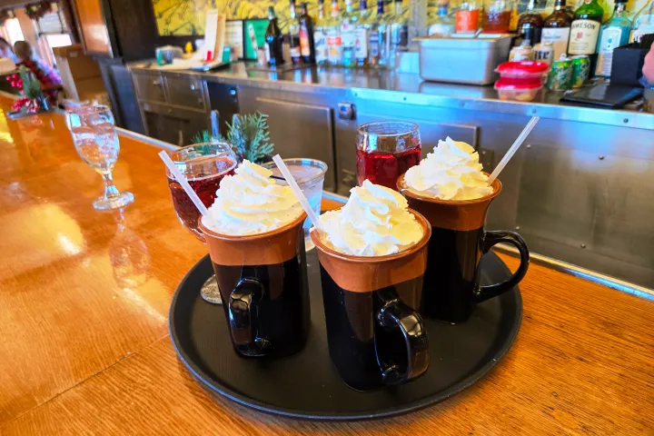 Tray with three mugs of whipped cream coffee and two drinks on a bar counter.