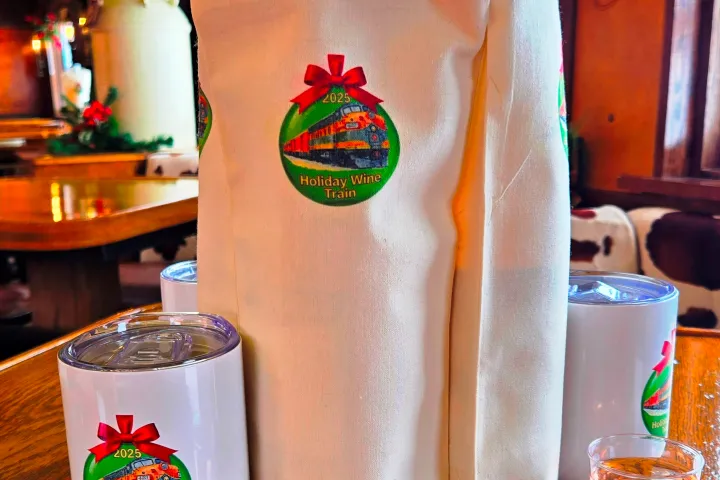 Wine bottles in a canvas bag with 'Holiday Wine Train' logo, cups, and shots on a wooden table.